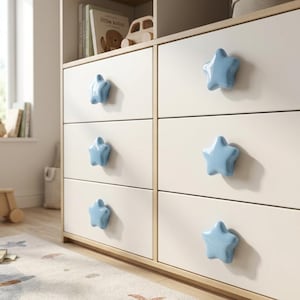 Op de afbeelding: Een ladekast van licht hout met zes lades, elk voorzien van een glanzende, lichtblauwe, stervormige knop. De ladekast heeft een strakke, gladde afwerking en de stervormige handgrepen zijn uniform in kleur en grootte over alle lades.