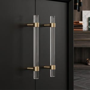 May include: Two vertical cabinet pulls featuring clear, cylindrical acrylic bars mounted with brushed brass metal hardware. Each handle is attached to a matte black cabinet door with two circular brass brackets, creating a clean, modern look.