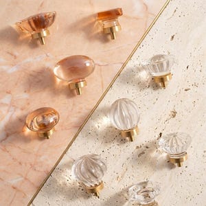 May include: A collection of cabinet knobs featuring clear and amber-tinted glass tops with brass-toned metal bases. The knobs vary in shape, including smooth oval, round, and ribbed designs, all mounted on a split-surface background.