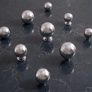 Puede incluir: Un conjunto de nueve pomos esféricos para armarios con acabado en cromo pulido. Cada pomo presenta una cabeza redonda, lisa y reflectante, unida a una pequeña base cilíndrica, dispuestos de forma dispersa sobre la superficie gris oscura.