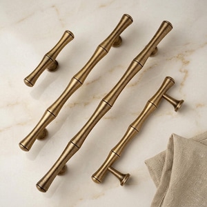 May include: Four brass-toned metal cabinet pulls with a textured bamboo-inspired design. The set includes one small knob-style pull and three elongated bar pulls of varying lengths, all featuring a warm, brushed metallic finish and rounded ends.