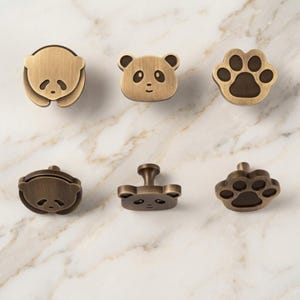 May include: A set of six metal cabinet knobs in a brushed brass finish. The designs include a curled panda, a panda face, and a paw print. Each knob features a central mounting post on the back, showcasing the front and side profiles of the hardware.