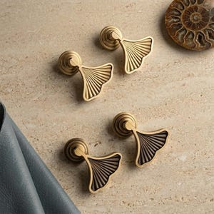 May include: Four metal cabinet knobs shaped like ginkgo leaves. Two knobs have a bright gold finish, and two knobs have a dark bronze finish. Each knob features a circular base and a fan-shaped handle with vertical ridges radiating from the center.