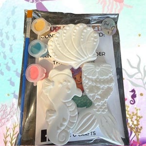May include: A craft kit in a clear plastic bag containing three white plaster figures shaped like a seashell, a seahorse, and a mermaid tail. The kit includes a paintbrush, three small paint pots in yellow, blue, and pink, and an instruction sheet.