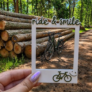 May include: A white rectangular photo frame featuring the text ride & smile in dark green script at the top and a dark green bicycle icon with a small smiley face at the bottom. The frame is held up to display a view of a bicycle in a forest.
