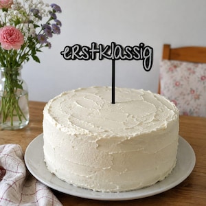 Peut inclure: Un gâteau rond recouvert de glaçage blanc sur une assiette blanche, surmonté d'une décoration de gâteau noire portant le mot "erstklassig" dans une police cursive stylisée. La décoration possède une tige verticale insérée au centre.