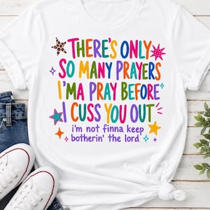 Może przedstawiać: Biała koszulka z krótkim rękawem z kolorowym napisem: There's Only So Many Prayers I'ma Pray Before I Cuss You Out, i'm not finna keep botherin' the lord. Wzór zawiera różne małe gwiazdki i błyszczące ikony w jasnych, żywych kolorach.