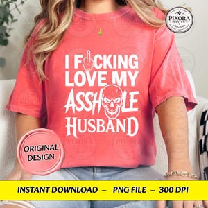 May include: A coral-colored short-sleeve t-shirt featuring white text that reads I FUCKING LOVE MY ASSHOLE HUSBAND, with a graphic of a middle finger replacing the letter U and a skull illustration replacing the letter O in the word ASSHOLE.