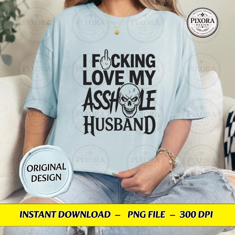 May include: A light blue t-shirt featuring black text that reads I F*CKING LOVE MY ASSHOLE HUSBAND, with a middle finger icon replacing the letter U and a skull graphic replacing the letter O in the word ASSHOLE, printed on the front.