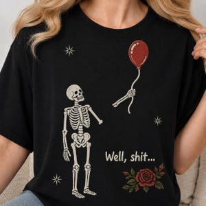 May include: A black t-shirt featuring a graphic of a skeleton looking up at a detached skeletal hand holding a red balloon. The design includes the text "Well, shit..." in a light-colored font, small white star accents, and a red rose with green leaves.