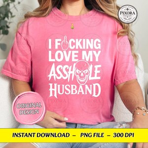 May include: A bright pink short-sleeved t-shirt featuring white text that reads I FUCKING LOVE MY ASSHOLE HUSBAND, with a graphic of a middle finger replacing the letter U and a skull illustration replacing the letter O in the word ASSHOLE.