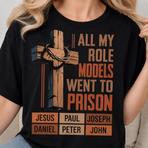 May include: A black t-shirt featuring a graphic of a wooden cross with a crown of thorns. The text reads: ALL MY ROLE MODELS WENT TO PRISON. Below, six rectangular boxes list the names: JESUS, PAUL, JOSEPH, DANIEL, PETER, and JOHN in orange text.