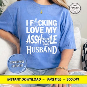 May include: A periwinkle blue short-sleeved t-shirt featuring white text that reads I FUCKING LOVE MY ASSHOLE HUSBAND, with a graphic of a middle finger replacing the letter U and a skull illustration replacing the letter O in the word ASSHOLE.