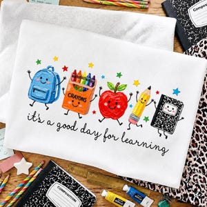 May include: A white t-shirt featuring a colorful graphic of a blue backpack, an orange crayon box, a red apple, a yellow pencil, and a black composition notebook with faces, accompanied by the text "it's a good day for learning".