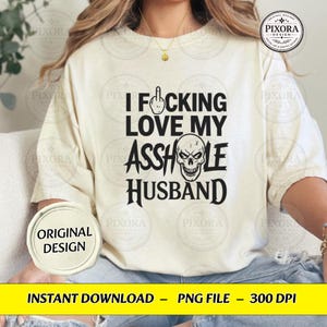May include: A cream-colored short-sleeve t-shirt featuring black text that reads I FUCKING LOVE MY ASSHOLE HUSBAND. The design includes a graphic of a hand making a middle finger gesture and a skull illustration replacing the letter O in asshole.