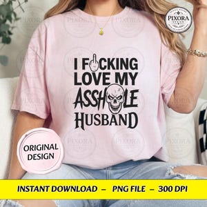May include: A light pink short-sleeve t-shirt featuring black text that reads I F*CKING LOVE MY ASSHOLE HUSBAND with a middle finger icon replacing the letter U and a skull graphic replacing the letter O in the word ASSHOLE.