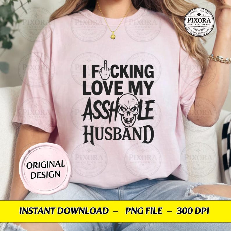 May include: A light pink short-sleeve t-shirt featuring black text that reads I F*CKING LOVE MY ASSHOLE HUSBAND with a middle finger icon replacing the letter U and a skull graphic replacing the letter O in the word ASSHOLE.