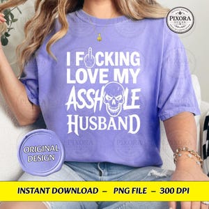 May include: A light purple short-sleeve t-shirt featuring white text that reads I FOCKING LOVE MY ASSHOLE HUSBAND, with a middle finger icon replacing the O in FOCKING and a white skull graphic integrated into the word ASSHOLE.