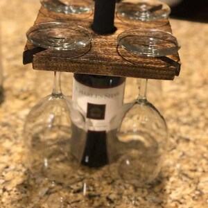 May include: A dark brown stained wooden wine glass holder with a square top featuring four slots for hanging stemware. The holder is positioned over a bottle of Charles Shaw wine, with the bottle neck extending through the center hole of the wood.