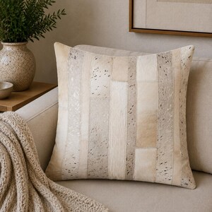 May include: A square throw pillow featuring a patchwork design of vertical strips in varying shades of cream, beige, and light gray. The surface is composed of textured animal hide panels with natural speckled patterns and smooth solid sections.