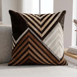 Könnte beinhalten: Ein quadratisches Zierkissen mit einem geometrischen Chevron-Muster, das aus genähten Paneelen in den Farben Dunkelbraun, Mittelbraun, Hellbraun und Cremeweiß besteht. Die Oberfläche hat eine texturierte Felloptik mit abwechselnden Diagonalstreifen.