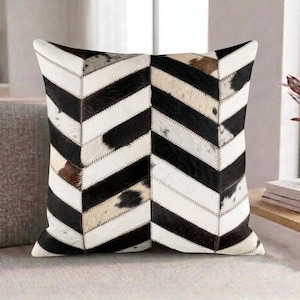May include: A square throw pillow featuring a chevron pattern made from stitched panels of black, white, and brown cowhide. The hair-on-hide material displays natural variations in color and texture, arranged in a symmetrical V-shaped design.