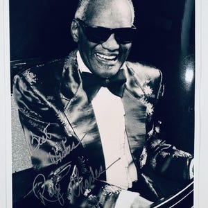 May include: A black and white photograph of Ray Charles wearing a dark patterned blazer with a white shirt and black bow tie, dark sunglasses, and a smiling expression. The image features a handwritten signature that reads Best Wishes Ray Charles.