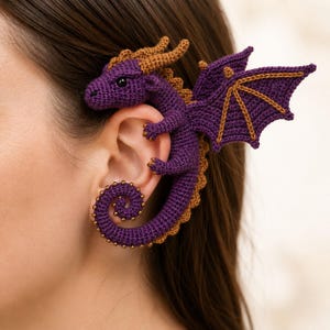 May include: A purple and brown crocheted dragon ear cuff. The dragon features a detailed head with small horns, a long body that curves around the earlobe, and a large wing extending outward. The design includes small gold-colored beads along the tail.