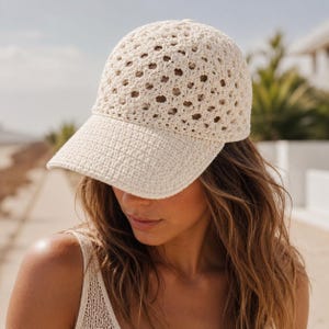 May include: A cream-colored baseball cap featuring an intricate, open-work crochet pattern across the crown and a solid, tightly stitched brim. The cap is constructed from yarn and displays a textured, geometric design throughout the dome.