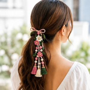 May include: A hair accessory featuring a crocheted dark green base with a pink bow, white and pink flowers, and dangling strands adorned with small beads and three tassels in shades of pink, cream, and dark green, secured in a ponytail.