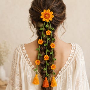 May include: A crochet hair accessory featuring a large orange sunflower at the top with three long green vine-like strands hanging down. Each strand is decorated with smaller orange flowers and leaves, ending in a solid orange yarn tassel.