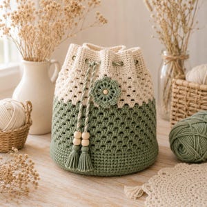 May include: A two-toned crochet bucket bag featuring a cream-colored upper section and a sage green lower section. The bag includes a drawstring closure with two sage green tassels, wooden beads, and a decorative crocheted flower accent.