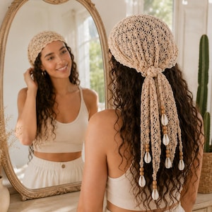 May include: A beige crochet headscarf featuring an intricate floral pattern, tied at the back with long, dangling ends. The ends are decorated with small, round wooden beads and white cowrie shells, creating a textured, bohemian-inspired accessory.