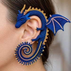 Puede incluir: Un pendiente tipo ear cuff de ganchillo con forma de dragón azul y detalles dorados en la columna, las alas y la cola. La cabeza del dragón descansa cerca de la parte superior de la oreja, y su larga cola en espiral tiene pequeñas cuentas doradas.