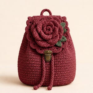 May include: A deep burgundy crochet backpack featuring a large, dimensional rose motif on the front flap. The bag includes a metallic bronze-toned push-lock closure, two braided drawstring cords, and small green crocheted leaves accenting the rose.