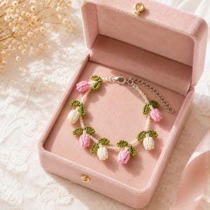 May include: A delicate bracelet featuring crocheted pink and white tulip flowers with green leaves attached to a thin white cord. The bracelet includes a silver-toned metal lobster claw clasp and an adjustable chain link extender for sizing.