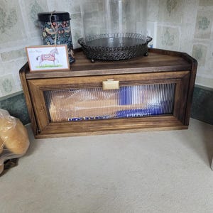 May include: A dark brown wooden bread box with a rectangular, fluted glass front panel and a gold-toned handle. The box features a hinged door and a flat top surface, providing storage for kitchen items while maintaining a visible interior.