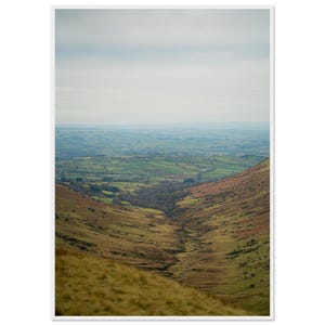 May include: A landscape view of a valley with brown, grassy slopes in the foreground leading down to a green, patchwork countryside under a cloudy, overcast sky. A small stream winds through the centre of the valley floor toward the horizon.