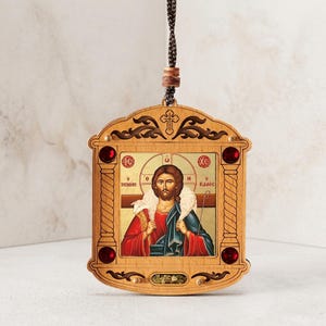 May include: A wooden hanging ornament featuring a central icon of a figure with a sheep draped over their shoulders. The frame is decorated with four dark red circular gems, carved scrollwork, a small cross, and a brown braided hanging cord.