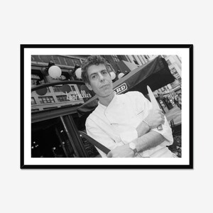 Può includere: Una fotografia in bianco e nero di uno chef con una giacca bianca a doppio petto e le braccia incrociate, che tiene in mano un grande coltello da cucina. Lo chef si trova davanti a un edificio con l'insegna Les Halles, in una cornice nera.