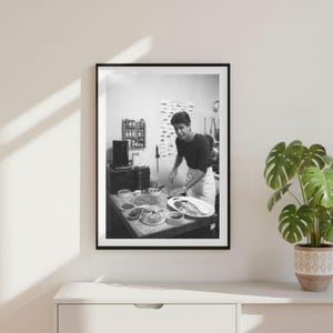 May include: A black and white framed photograph depicting a person in a dark long-sleeved shirt and apron standing in a kitchen. The person is preparing food on a counter filled with small bowls, a microwave, a spice rack, and a fish chart.