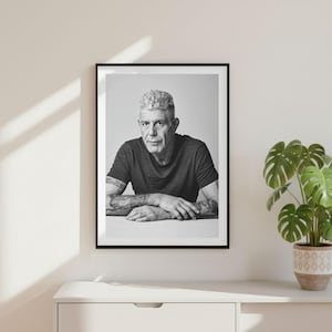 May include: A black and white portrait print of Anthony Bourdain in a black t-shirt, featuring visible arm tattoos, displayed in a thin black frame. The subject is shown from the chest up, resting arms on a surface, looking directly at the camera.