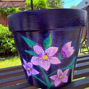 May include: A dark blue tapered plant pot featuring a hand-painted floral design. The artwork displays three light purple flowers with yellow centers and green leaves, set against a textured, deep navy blue background with visible brushstrokes.