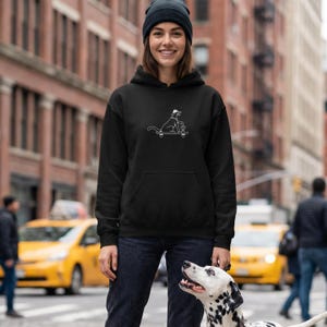 Puede incluir: Una sudadera negra con capucha que presenta un gráfico de líneas blancas de un perro con sombrero sobre un monopatín sosteniendo una taza. La sudadera tiene mangas largas, un bolsillo tipo canguro y un gorro negro a juego en la cabeza.