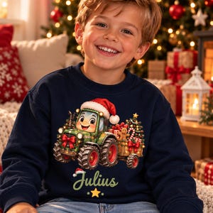 Könnte beinhalten: Ein dunkelblaues Langarm-Sweatshirt mit einer Grafik eines grünen Traktors mit Weihnachtsmannmütze, der einen Anhänger voller Weihnachtsgeschenke, einen Baum und einen Lebkuchenmann zieht, mit dem Namen Julius in hellgrüner Schrift darunter.