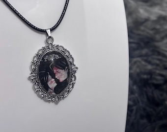 Puede incluir: Un collar con colgante ovalado que presenta un marco de filigrana en tono plateado y un fondo negro con una ilustración de dos figuras frente a frente. El texto "FoMa" está impreso en la parte superior. El colgante cuelga de un cordón negro.