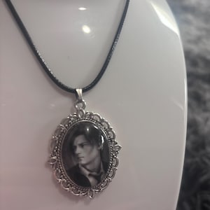 Może przedstawiać: Srebrzysty naszyjnik z owalną zawieszką z misterną filigranową ramką otaczającą czarno-biały portret osoby z krótkimi włosami. Zawieszka wisi na prostym czarnym sznurku, tworząc wyraźny kontrast pomiędzy metalem a sznurkiem w tym naszyjniku.