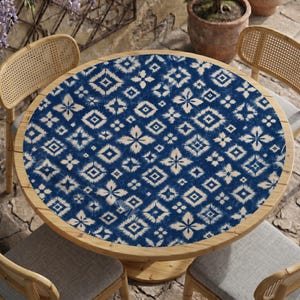 May include: A round table cover featuring a dark blue and off-white ikat-style geometric pattern with repeating diamond and floral shapes. The fabric is laid flat across the circular wooden tabletop, displaying a consistent, symmetrical print.
