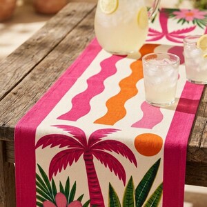May include: A rectangular table runner featuring a cream-colored center with a vibrant pattern of pink palm trees, orange suns, green tropical leaves, and pink flowers, bordered by solid pink stripes and accented with wavy orange and pink lines.