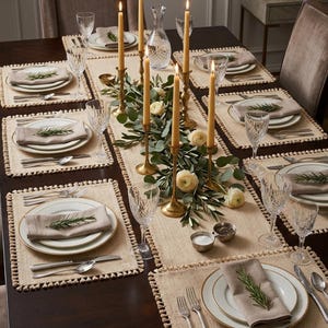 Puede incluir: Una mesa de comedor con seis servicios que incluyen manteles individuales beige con borde de pompones, platos blancos con borde dorado, servilletas beige y copas de cristal. Un camino de mesa central sostiene candelabros de latón.